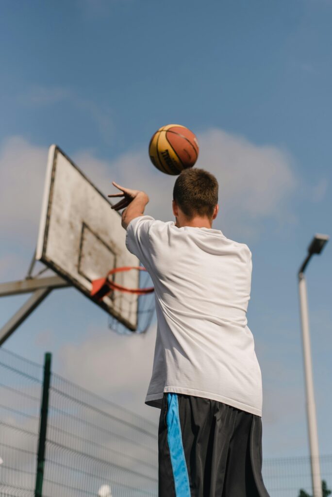 Back view of a young man making a basketball shot on an outdoor court under clear skies.